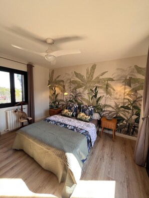 Une chambre avec un lit, un ventilateur de plafond, un radiateur, une fenêtre avec des rideaux et un mur orné d’une fresque représentant des plantes tropicales.