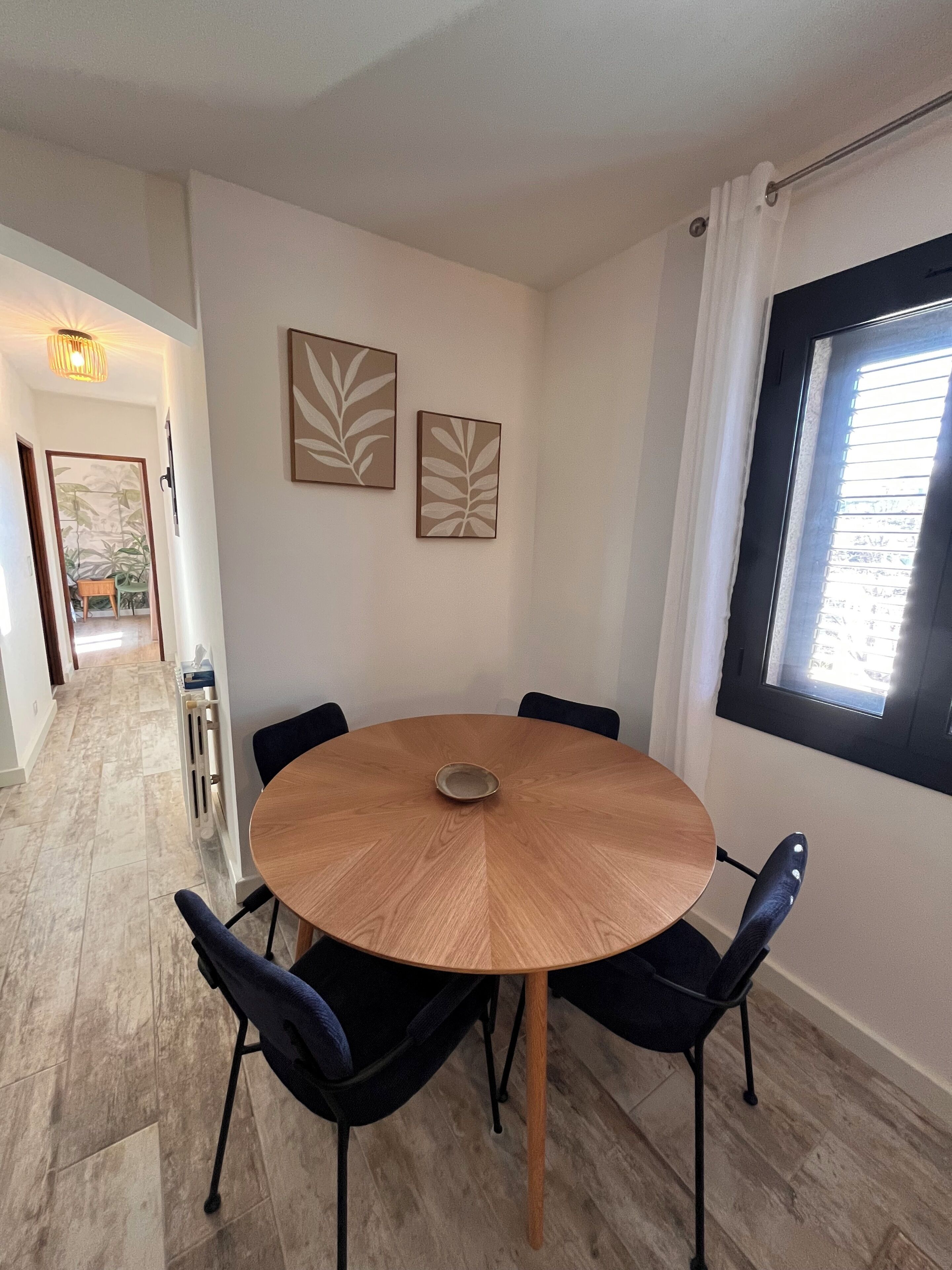 A round wooden table with four chairs, two wall-mounted artworks, a window with blinds, and a hallway leading to another room.