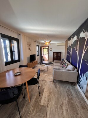 Un salon comprenant un canapé, une table à manger avec des chaises, un téléviseur, un ventilateur de plafond et une grande fresque murale.
