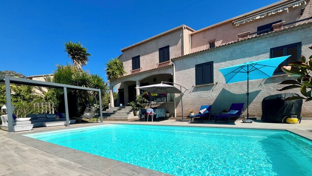 A swimming pool with lounge chairs and umbrellas, a covered seating area, and a building with a stucco exterior.