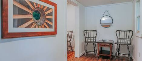 A hallway with wooden flooring, a framed artwork, two chairs, a small table, and a mirror on the wall.
