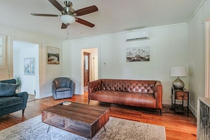 Una sala de estar con un sofá de cuero marrón, un sillón gris oscuro, una mesa de centro de madera y un ventilador de techo.