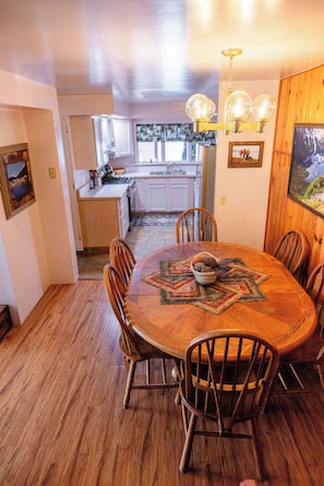 Un comedor con una mesa de madera, sillas y una araña. Se observa una cocina al fondo con armarios blancos y un refrigerador.