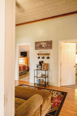 A room with a wooden floor, a sofa, a coffee table, a TV, a bed, a wall-mounted shelf with mugs, and a door.