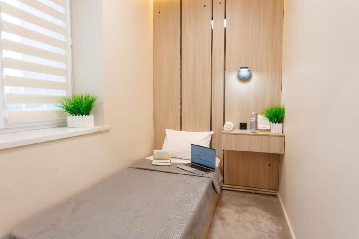 A small bedroom with a bed, a laptop, a bedside table with a lamp, and a window with blinds.