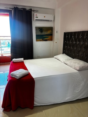 Habitación de hotel con cama, una toalla roja, un cuadro de una playa y un equipo de aire acondicionado.