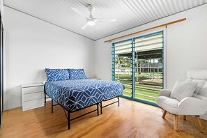 A bedroom with a bed, a white chair, a ceiling fan, and a large window with blinds. - Fish Tales - Absolute Waterfront Toogoom-Sleeps 8-Air Con-Wi-Fi-Pets (Toogoom)