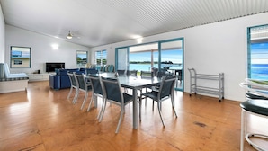 Ein geräumiges Zimmer mit einem großen Esstisch, Stühlen, einer Couch und einem Fernseher. Große Fenster bieten Blick aufs Meer.