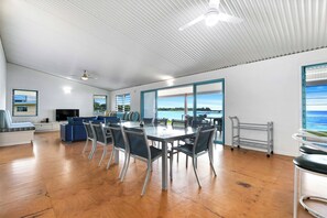 Ein geräumiges Zimmer mit einem großen Esstisch, Stühlen, einer Couch und einem Fernseher. Große Fenster bieten Blick aufs Meer.