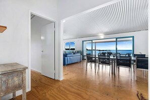 Ein geräumiges Zimmer mit Holzboden, einem Essbereich mit Stühlen und Tisch, einem blauen Sofa und Blick durch große Fenster.