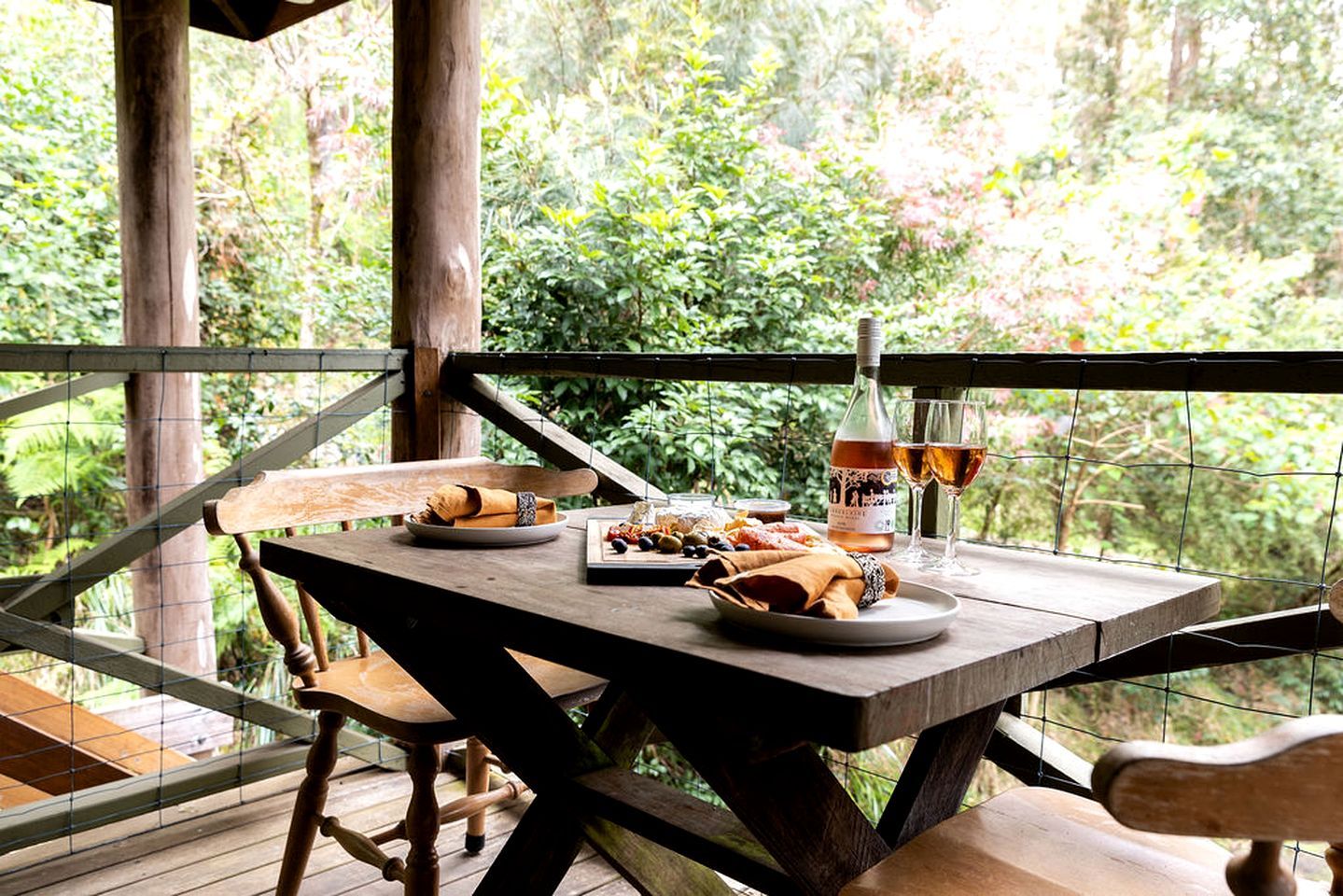 An outdoor wooden table set with food and drinks, surrounded by a wooden fence and lush greenery.