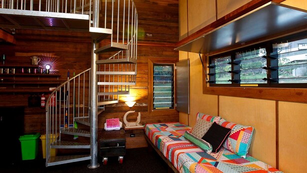 A cabin interior with a spiral staircase, a sofa with a colorful quilt, and a window with blinds. - Gorgeous Queensland Tree House with Great Views Perfect for Glamping, Gold Coast (Gold Coast)