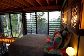 A bedroom with a large bed, a view of trees through the window, and a wooden ceiling.