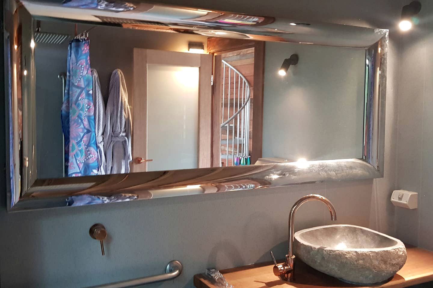 A bathroom with a large mirror, a stone sink, a towel rack with towels, a handrail, and a door leading to another room.