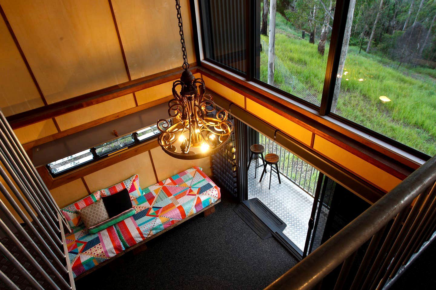 A room with a colorful quilted bench, a hanging light fixture, and a view of a grassy outdoor area through large windows.