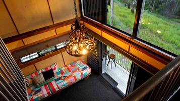 A room with a colorful quilted bench, a hanging light fixture, and a view of a grassy outdoor area through large windows.