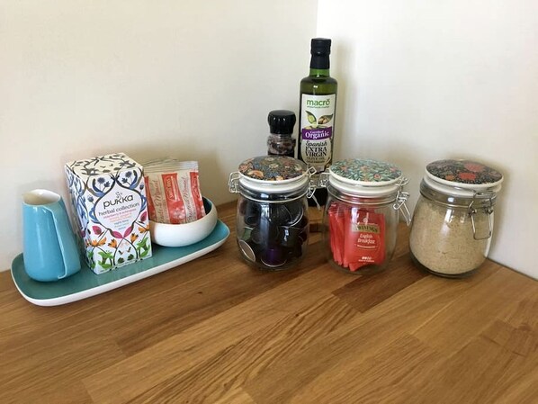A wooden table with various jars and containers, including a box labeled 'Purka herbal collection,' a bottle of 'macró Organic Spanish EXTRA VIRGIN OLIVE OIL,' and several spice jars.