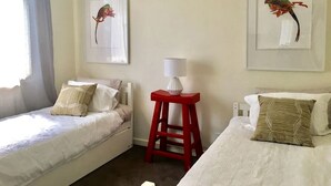 Une chambre avec deux lits simples, un tabouret rouge et deux tableaux encadrés accrochés au mur.