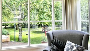Ett rum med ett stort fönster, en grå fåtölj med en kudde, ett soffbord i glas och ett litet sidobord i rotting.