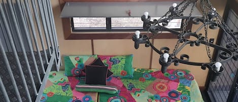 Una habitación con estructura metálica para la cama, una colcha colorida, una ventana con persianas y una lámpara colgante.