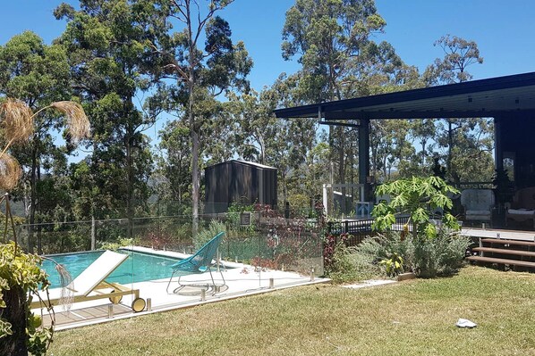 Un jardin avec une piscine, un patio couvert et un espace clos bordé d’arbres.