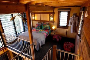 A cozy cabin-style bedroom with a bed, colorful pillows, a hanging light fixture, and a window with clothes hanging outside.