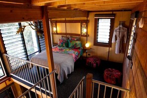 A cozy cabin-style bedroom with a bed, colorful pillows, a hanging light fixture, and a window with clothes hanging outside.