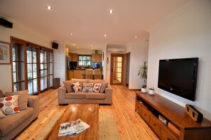 A living room with a sofa, a coffee table with magazines, a TV, and a kitchen in the background.