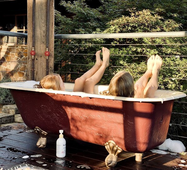 A vintage bathtub with two people reclining inside, surrounded by a wooden deck and greenery.