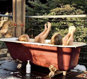 A vintage bathtub with two people reclining inside, surrounded by a wooden deck and greenery.