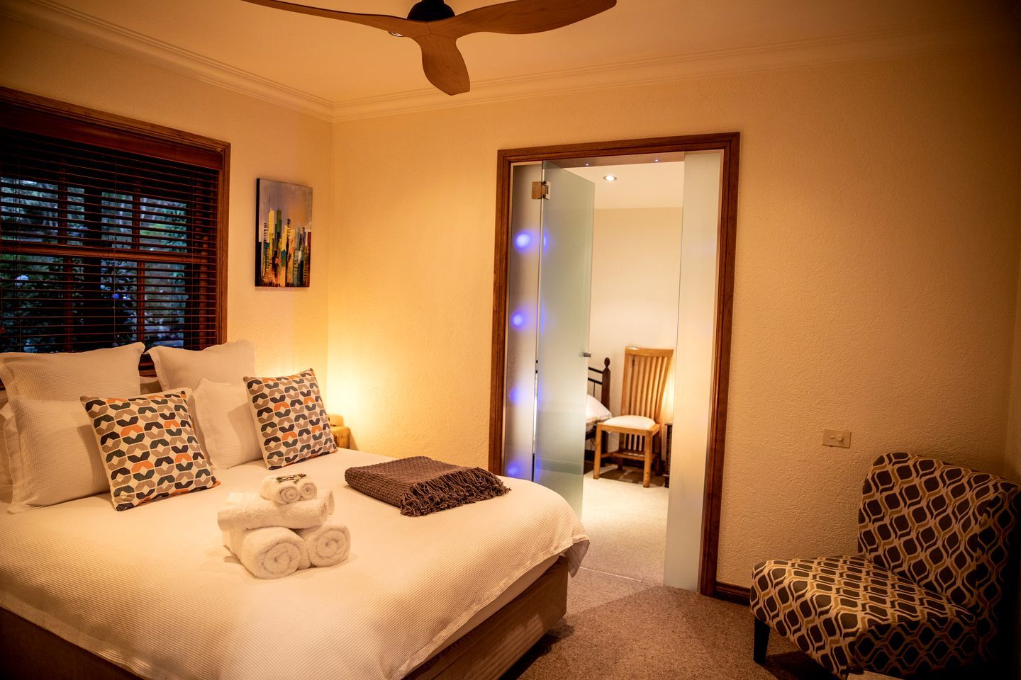 A bedroom with a bed, pillows, a chair, a painting, and a ceiling fan.