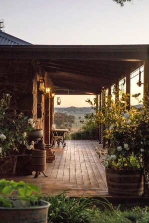 A wooden deck leading to a covered outdoor area with rustic stone walls, potted plants, and a view of the countryside.