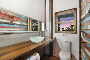 Ein Badezimmer mit einem Holzkosmetikschrank, einem Glaskonstruktionsbecken, einer Toilette und einem Fenster mit Sonnenuntergangsblick.