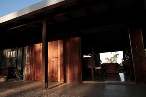 Un espacio al aire libre cubierto con suelo de hormigón, pilares de madera y una puerta que da al interior.
