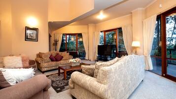 A living room with a sofa, armchairs, a coffee table, and a TV. There are large windows with curtains, a fireplace, and a framed picture on the wall.
