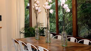 A dining table with chairs, three glass vases with branches and white flowers, and a window with a view of greenery.