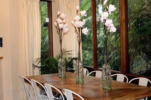 A dining table with chairs, three glass vases with branches and white flowers, and a window with a view of greenery.