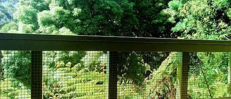 A white cup of coffee on a wooden table with a view of green trees through a mesh fence.