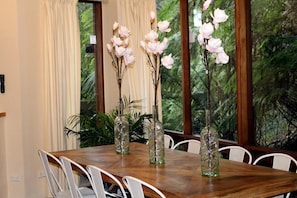 Une table à manger avec des chaises, trois vases en verre ornés de branches et de fleurs blanches, et une fenêtre donnant sur un paysage verdoyant.