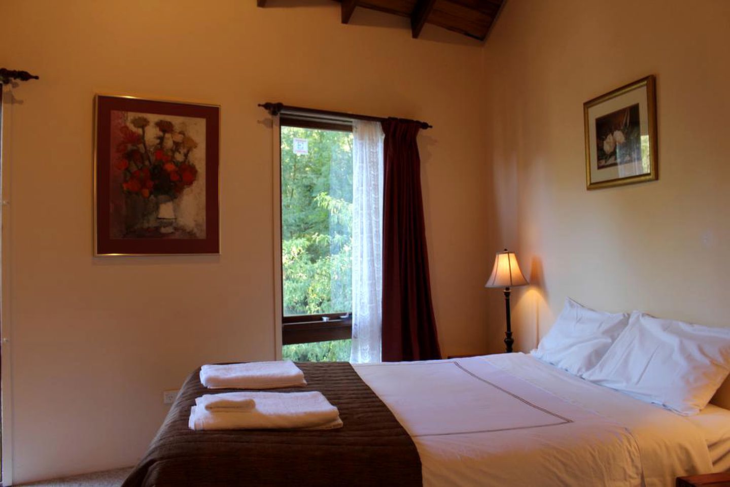 Une chambre d’hôtel avec un lit, deux serviettes, une table de chevet avec une lampe, une fenêtre avec des rideaux et deux tableaux encadrés accrochés aux murs.