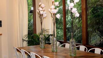 A dining table with chairs, three glass vases with branches and white flowers, and a window with a view of greenery.
