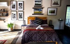 Quarto bem organizado com cama, mesas de cabeceira, planta em vaso e quadros nas paredes.