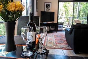A table with a wine bottle and two glasses, a vase with yellow flowers, and a view of a living room with a sofa and a TV. - Marvelous Halls Gap Cabin Ideal for Romantic Getaways in Victoria (Halls Gap)