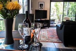 Et bord med en vinflaske og to glas, en vase med gule blomster, og udsigt til en stue med en sofa og et fjernsyn.