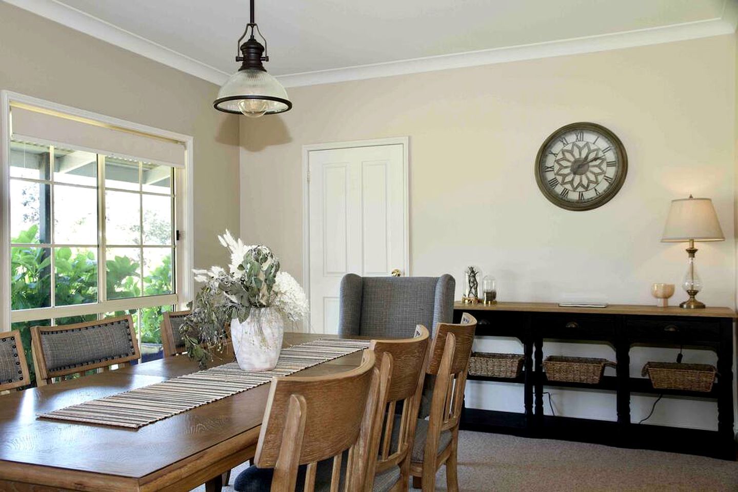 A dining area with a wooden table, chairs, a vase with flowers, a wall clock, and a lamp.