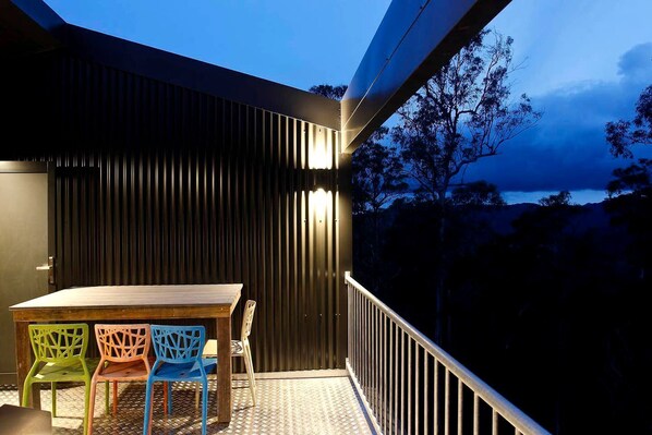 A balcony with a table and chairs, a view of trees and a body of water, and a building with vertical siding.