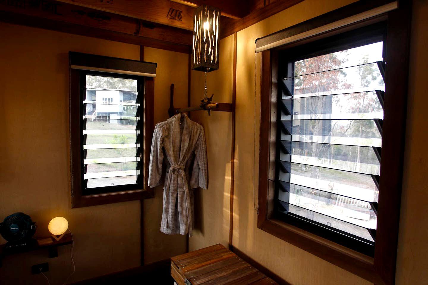 A room with a wooden bench, a robe hanging on a rack, a lamp, and two windows with blinds.