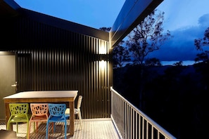Un balcon avec une table et des chaises, une vue sur des arbres et une étendue d’eau, et un bâtiment dont le revêtement extérieur est composé de panneaux verticaux.