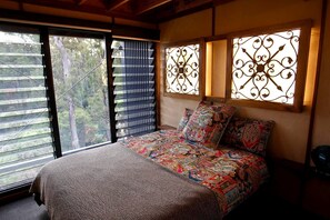 A bedroom with a large bed, patterned bedding, a view through a window with blinds, and decorative ironwork on the wall.