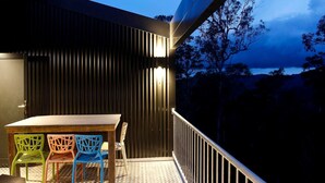 Un balcón con mesa y sillas, vistas a árboles y una masa de agua, y un edificio con revestimiento vertical.